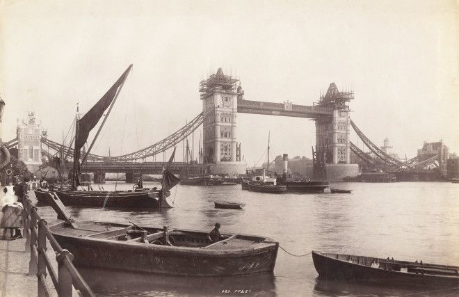 The Tower Bridge is seen under construction with river traffic in the foreground.